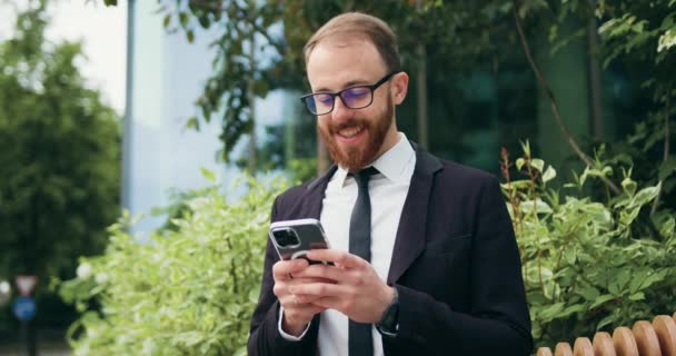 Νεαρός Χαμογελαστός Επιχειρηματίας Χρησιμοποιεί Ένα Smartphone Ενώ Κάθεται Ένα Παγκάκι — Αρχείο Βίντεο