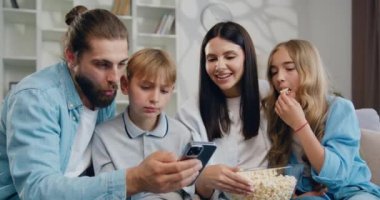 Mutlu, genç bir aile kanepeye uzanıyor. Sevimli baba kız ve oğul anne eli ile birlikte oturma odasında akıllı telefon uygulaması kullanıyorlar. Aile akıllı telefon bağımlılığı. Yavaş çekim.