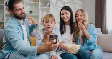 Şirin okul çocukları olan mutlu aile ebeveynleri kanepeye oturup komik akıllı telefonlar kullanarak konferans konuşmaları yapıyor, çevrimiçi içeriği izliyor, gülümsüyor, gülüyor. Yavaş çekim.