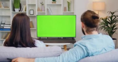 Mutlu çift televizyon izliyor ve kanalları değiştiriyor. Modern oturma odasındaki yeşil ekrana bakıyor. Kadın ve erkek krom televizyon izliyor. Yavaş çekim.