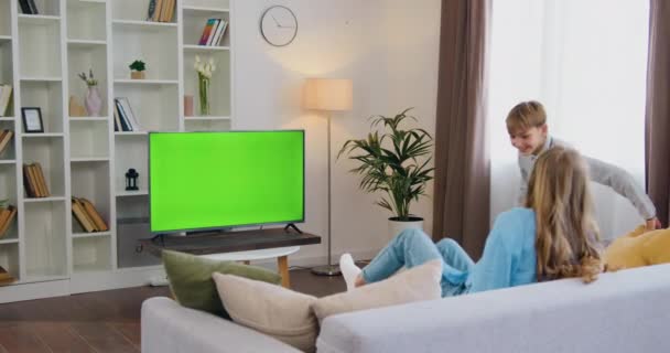 年轻的父亲 儿子和女儿坐在沙发上 笑着看绿屏电视 美丽的家庭正在客厅里用彩色钥匙看电视 慢动作 — 图库视频影像