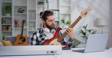 Modern sınıfın masasında online elektro gitar dersi alan yakışıklı sakallı adam. Genç müzisyen, modern işyerinde dizüstü bilgisayar ve elektro gitar kullanarak yeni bir proje üzerinde çalışıyor.
