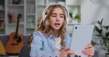 Beyaz kulaklıklı, uzun kıvırcık saçlı, müzik dinleyen ve masada otururken dijital tabletle şarkı söyleyen güzel bir kızın portresi. Yavaş çekim