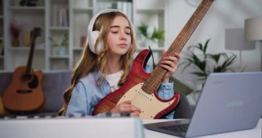 Kulaklık takıp evde gitar çalmayı öğrenen güzel genç kız dizüstü bilgisayardaki dersleri kullanıyor. Uzun saçlı güzel bir kız dizüstü bilgisayarla evde gitar çalıyor. Yavaş çekim