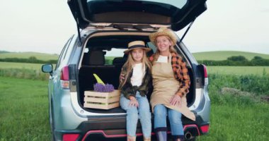 Çiftçi aile annesi ve kızı, önlük ve şapka giyip gün batımında bir tarlada kameraya bakan taze lavanta ile ahşap kutunun yanındaki bir arabanın bagajında oturuyorlar. Aile işi konsepti