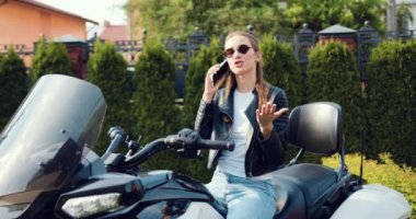 Yüksek atkuyruklu, siyah deri elbiseli şık bir kadın spor motosikletinin üzerinde otururken cep telefonuyla konuşuyor. Konuşan portre kızı Green Street 'te akıllı telefon kullanıyor. Yavaş