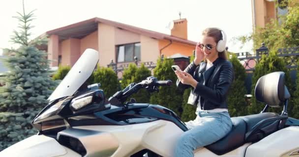 穿着时髦的白色大耳机坐在摩托车上 用智能手机欣赏音乐 有着优雅发型的令人毛骨悚然的女性形象 穿着优雅 令人毛骨悚然的漂亮女孩的户外肖像 — 图库视频影像