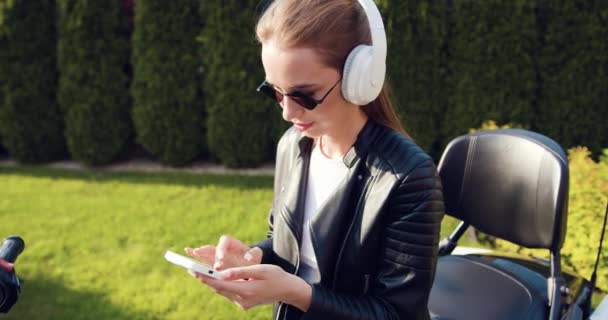 大きなホワイトヘッドフォンを着用している美しい少女は 携帯電話を使用し スポーツバイクに座っている間 幸せに音楽を聞きます スローモーション — ストック動画