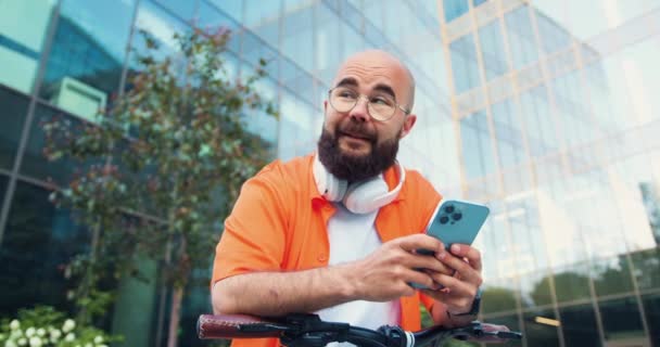 穿着眼镜站在户外骑自行车 用智能手机发短信 面带微笑的英俊年轻白人男子的画像 英俊的年轻男子 骑自行车 在电话上打字 慢动作 — 图库视频影像