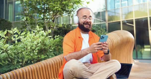 穿着橙色衬衫 戴着眼镜 头戴智能手机的英俊男子的画像 在社交网络上书写 并在网上发牢骚 他们坐在自己旁边的长椅上疯狂地聊天 — 图库视频影像