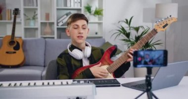 Yakışıklı ergen blogger video sınıfı özel dersini kaydediyor modern akıllı telefondan ev telefonunda gitar çalmak için. Genç çocuk internet işitme cihazı için elektro gitar çaldığını gösteriyor. Yavaş çekim