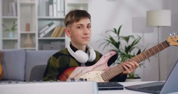 年轻英俊男孩的肖像 带着耳机和电子吉他 在灯光室或音乐学校看着相机 十几岁的男孩 学习弹奏乐器和看相机 慢动作 — 图库视频影像