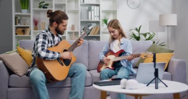 Genç müzisyen öğretmen ve 10 yaşındaki kız oturma odasında kanepede oturuyor, gitar çalıyor ve birlikte müzik dersinin tadını çıkarıyor, kapalı alanda. Yavaş çekim