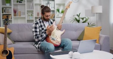 Yetenekli müzisyen, modern stüdyoda kanepede oturup gitar çalarak dizüstü bilgisayarda ders veriyor. Beyaz erkek müzisyen müzik aleti kullanıyor ve öğrencilere bilgisayar yoluyla video dersi veriyor.
