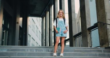 Neşeli kız okul binasının yanında sırt çantasıyla poz veren ve kameraya gülümseyen kitaplara sarılıyor. Modern eğitim, üniversite harcı ve burslar, yurtdışında öğrenim ve öğrenim. Yavaş çekim.