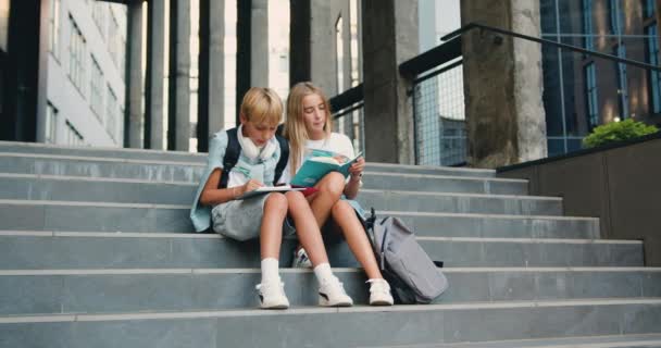 快乐的高中生坐在校园附近的楼梯街上 课前谈笑风生 白人男孩和女孩在课后一起在户外写书 — 图库视频影像