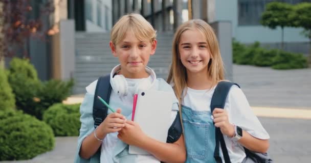 伟大的小学 快乐的男孩和女孩同学们 带着书本和背包 笑着看着一起在现代学校建筑附近的室外相机 青少年的学习和生活方式概念 — 图库视频影像