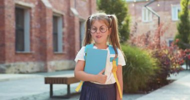 Gözlüklü, sırt çantalı ve kitaplı, kamera arkası okuluna bakan küçük tatlı bir kızın portresi. Eğitimli küçük çocuk. Mutlu öğrenci eğitimi. Yavaş çekim.