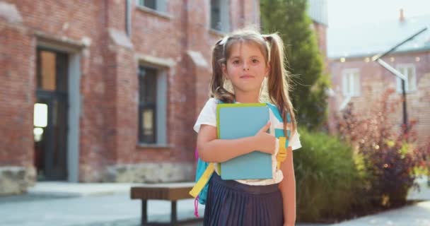 可爱的小学生小女孩拿着书本站在教学楼附近看着摄像机 孩子们把头靠在肖像上 回学校去慢动作 — 图库视频影像