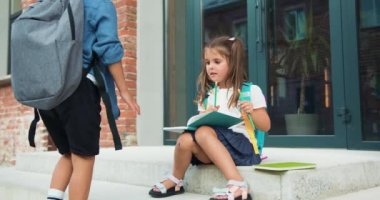 Küçük erkek arkadaş, küçük kıza gelip okul binasının arka planında otururken beşlik çakar. Konuşurken eğlenen bir çift. Çocuk okulun yanındaki planlamacıya yazı yazıyor. Öğrenci konsepti. Yavaş çekim.