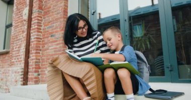 Güzel, gülümseyen genç bir kadın ve sırt çantalı mutlu küçük oğlu derslerden sonra okul binasının arka planında otururken not defterine bir şeyler yazıyorlar. Eğitici