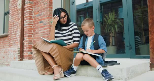 美丽的微笑的年轻女子和她快乐的小儿子 背着背包 在课后坐在学校大楼的楼梯上 用铅笔在笔记本上写下了一些东西 — 图库视频影像
