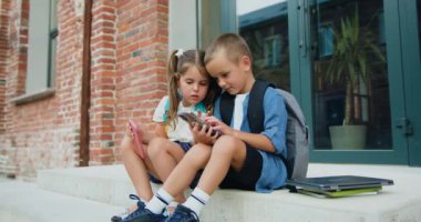Modern okul binasının yanındaki merdivenlerde oturan iki küçük öğrenci. Akıllı telefondan interneti arayan ve arkadaşına gösteren sevimli arkadaşlar. Öğrenme ve yaşam tarzı kavramı