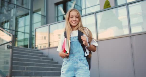 陽気な笑顔のかわいい白人女子高生 学校の背景でカメラにポーズする学校の近くの階段に立っているバックパック 小学生の肖像画 学校に戻る — ストック動画