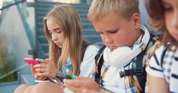 オンラインで通信する携帯電話を使用して屋外の階段に座っている子供たちのグループの近く 多様な子供たちは 晴れた日に街頭でリラックスしながら 携帯電話でインターネットを使用しています スローモーション — ストック動画
