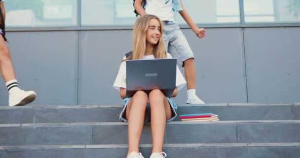 長髪の可愛い女の子はコンピュータデバイスを使用し 肯定的な多様な学校フレンドの屋外グループは女性の友人を迎えますが 女の子は階段に座ってラップを使用し 研究のために準備します — ストック動画