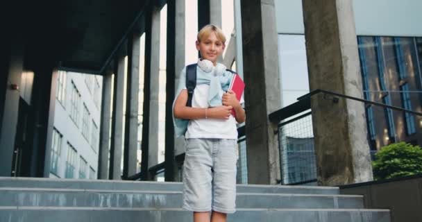 バックパックと屋外の学校の建物の背景にカメラの上に置かれた階段の上に立っている高価な見栄えの慎重なスクールボーイ 小学生の肖像画 スローモーション — ストック動画