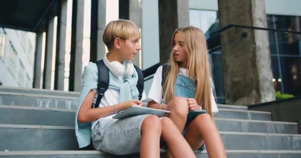 一个积极的青少年男孩和一个女孩坐在学校附近的楼梯上 玩的开心 谈论过去的讲座 在阳光明媚的夏日 在户外用石碑装饰 慢动作 — 图库视频影像