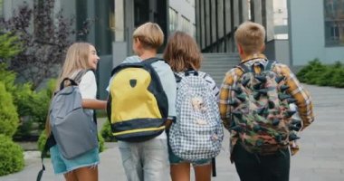 Renkli sırt çantalı dört akıllı sınıf arkadaşı okula giderken ve dışarıda eğlenerek konuşuyorlar. Arkadan bak. Yavaş çekim