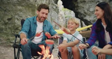 Genç sakallı baba ve yakışıklı oğul kamp ateşinde sosis pişiriyor. Aile tatili sırasında kaya yakınlarındaki ormanda konuşup gülüyorlar. Seyahat zamanı konsepti. Aile seyahati hayali konsepti