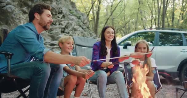 笑顔の家族の父親と息子のローストソーセージは 岩の自然の近くの森でピクニック中にキャンプファイヤーの上に 週末に2人の子供が森でピクニックをするハッピーな両親 女性の男と — ストック動画