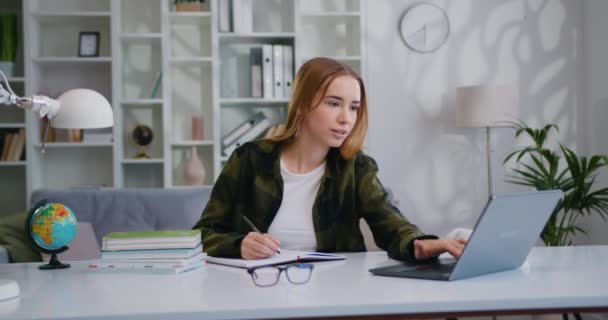 年轻的女商人坐在办公室的办公桌前 一边用笔记本电脑在笔记本上写字 年轻的女企业家计划工作日 组织会议 学生在抄本上记笔记 观看网络研讨会 — 图库视频影像