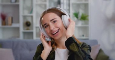Akıllı telefon ve kablosuz kulaklıkla müzik dinleyen, ışık odasında poz veren ve kameraya bakan güzel, gülen genç bir kadının yüzünü kapatın. Yavaş çekim