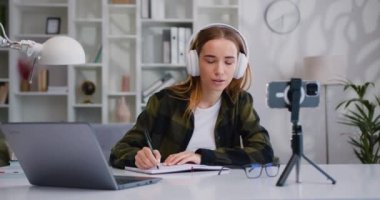 Kulaklıklı genç üniversiteli kız, online derste cep telefonuna bakarak masaya oturmuş notlar yazıyor. Sanal öğrenme kavramı, online eğitim. Yavaş çekim