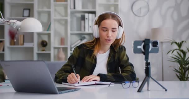 在网上课堂上 戴耳机的女大学生通过查看手机坐在桌边写笔记 虚拟学习的概念 在线教育 慢动作 — 图库视频影像