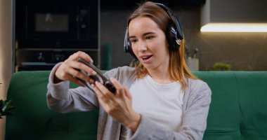 Kanepede oturan, kulaklıklı, cep telefonlu, rekabetçi video oyunu oynayan beyaz bir kadın. Akıllı telefonu, kulaklığı ve sosyal medya bağlantısı olan bir kadın oturma odasında video oyunu oynuyor