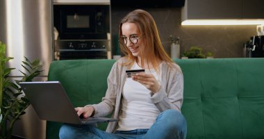 Güzel kız elinde banka kartı tutuyor ve evdeki modern dizüstü bilgisayarla online ödeme yapıyor..