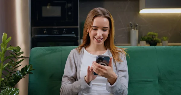 Mutfak arka planında yeşil bir koltukta otururken cep telefonu kullanan mutlu beyaz kadın. Sarı saçlı gülümseyen kız rahat koltukta dinleniyor ve akıllı telefondan internette geziniyor.