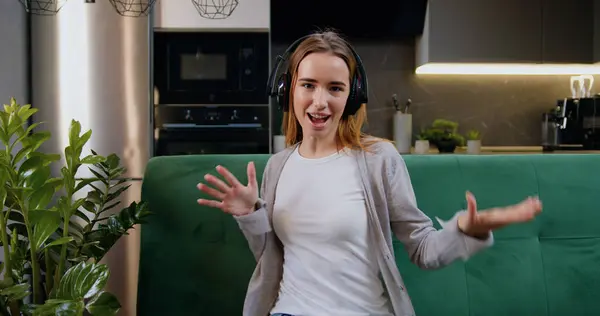 Kablosuz kulaklık takan neşeli genç bir kadın müzik dinliyor, kanepede dans ediyor, serbest dolaşımın tadını çıkarıyor. Kız ev mutfağının arka planında telefon dinleyerek şarkı söylüyor.