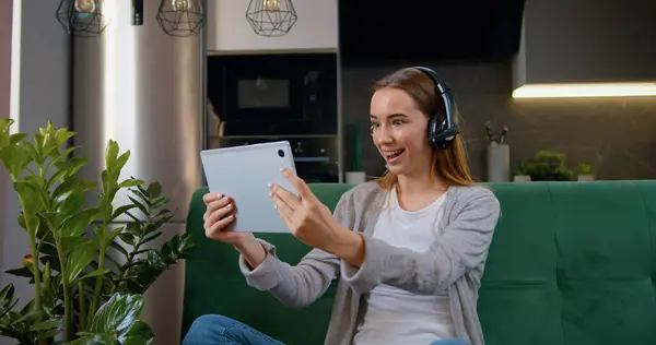Dijital tablet bilgisayarlı mutlu beyaz kız evdeki kanepede oturuyor. Gülümseyen kadın tabletle video çekiyor. Oturma odasındaki arkadaşlarıyla internet üzerinden iletişim kuruyor.
