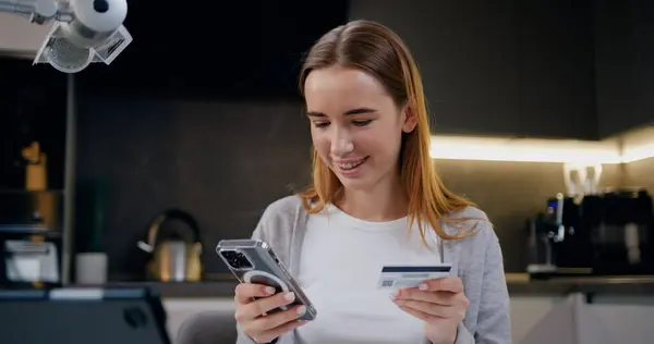 Beautiful girl entering credit card number for online shopping using mobile phone sitting at home kitchen. Smartphone communication technology banking money finance browsing and paying.