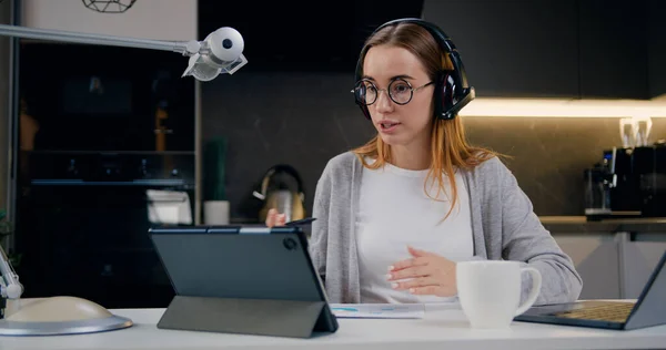 Freelancer female talking in a video conference on line with a headset, microphone and tablet device sitting while sitting the desk at home. Communication, video call online meeting with colleagues