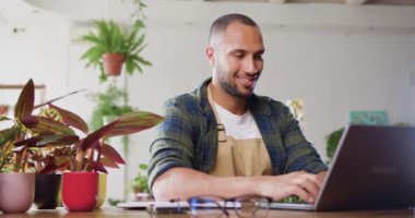 Owner male of flower shop working on laptop computer in workplace sitting in floral store. Man florist in apron looking at laptop and making sales order. Slow motion.