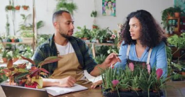 Küçük parlak çiçekçide dizüstü bilgisayar kullanan, daktilo yazan ve envanter çıkaran Afrikalı Amerikalı bir çiçekçi. Önlüklü kadın çalışan erkek meslektaşımla konuşuyor. Çiçekçi adam bitkileri kontrol ediyor ve kayıtları tutuyor.