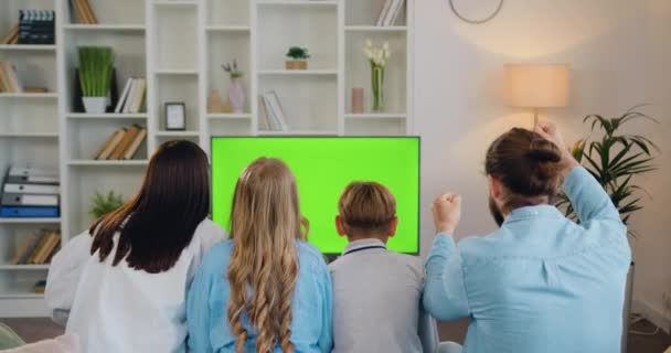 かわいい学校の子供たちがカウチに座って 緑色のクロマのキー画面を見て 自宅でリラックスしている幸せな家族の大人の両親 スポーツマッチのサポートを見ている居心地の良い部屋の男女と子供 — ストック動画