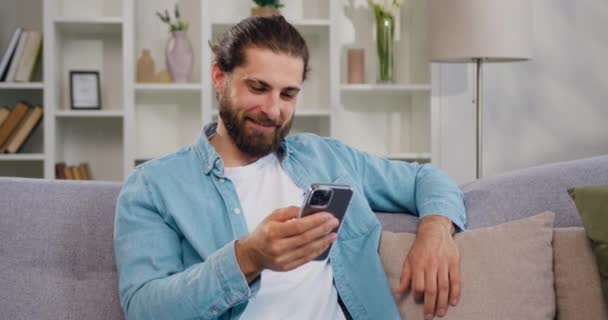 Latynos Uśmiechnięty Człowiek Zdalnie Pracuje Online Pomocą Smartfona Siedząc Kanapie — Wideo stockowe